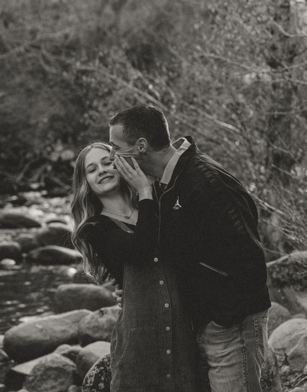 A couple shares joyful laughter while walking along a rocky stream in a natural outdoor setting.
