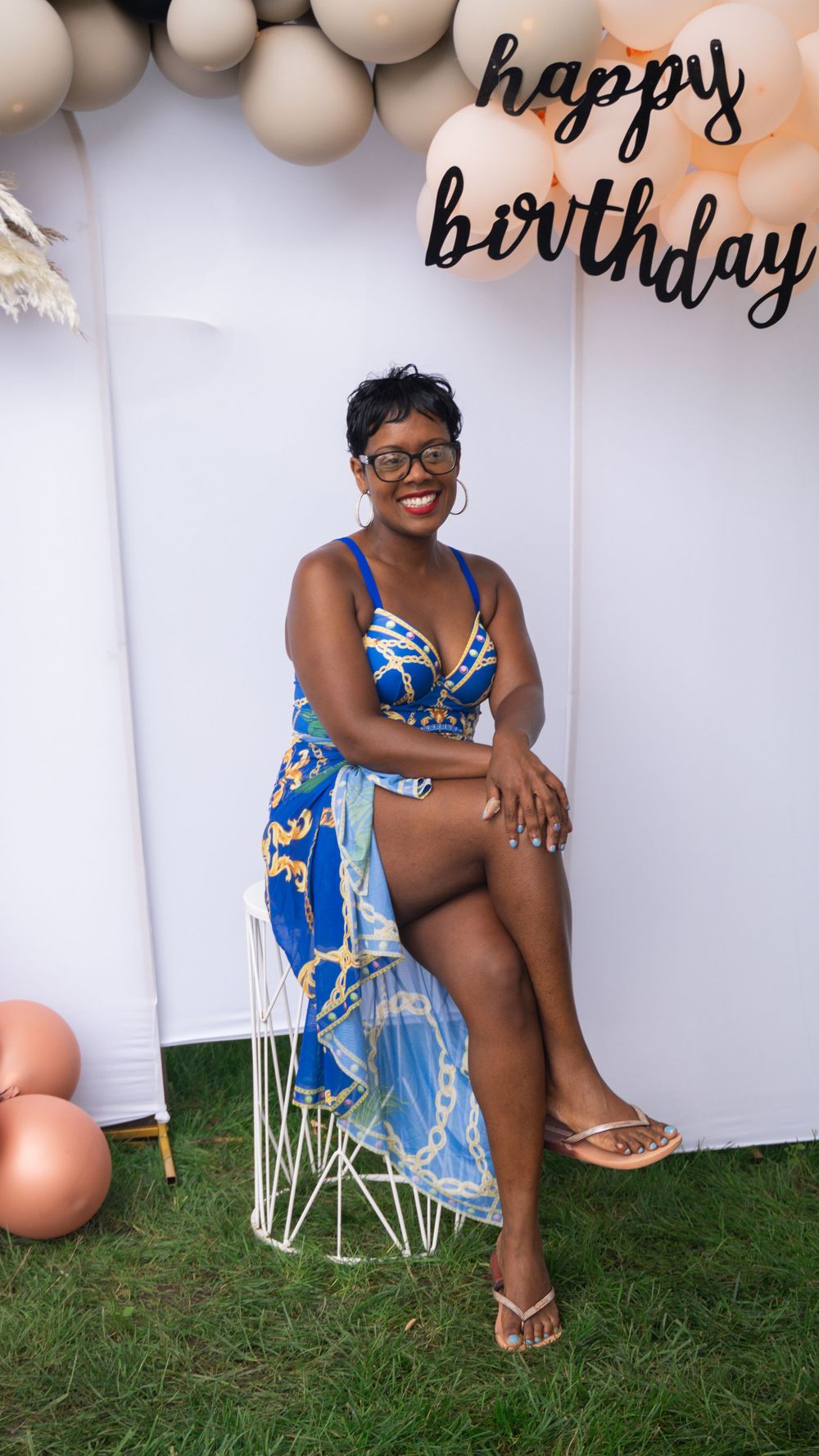 Birthday celebration backdrop with balloons and decorations featuring someone in a blue floral dress seated on a white chair.
