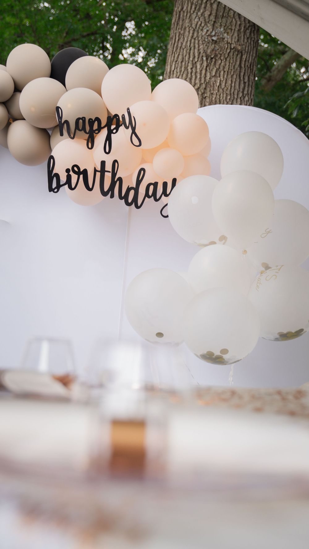 Elegant birthday party setup with neutral tone balloon arch featuring 'happy birthday' text against a natural outdoor backdrop.