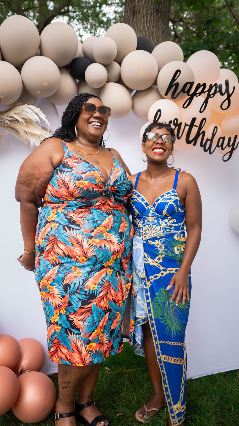 Two people wearing colorful tropical print dresses pose by a balloon arch at a birthday celebration outdoors.