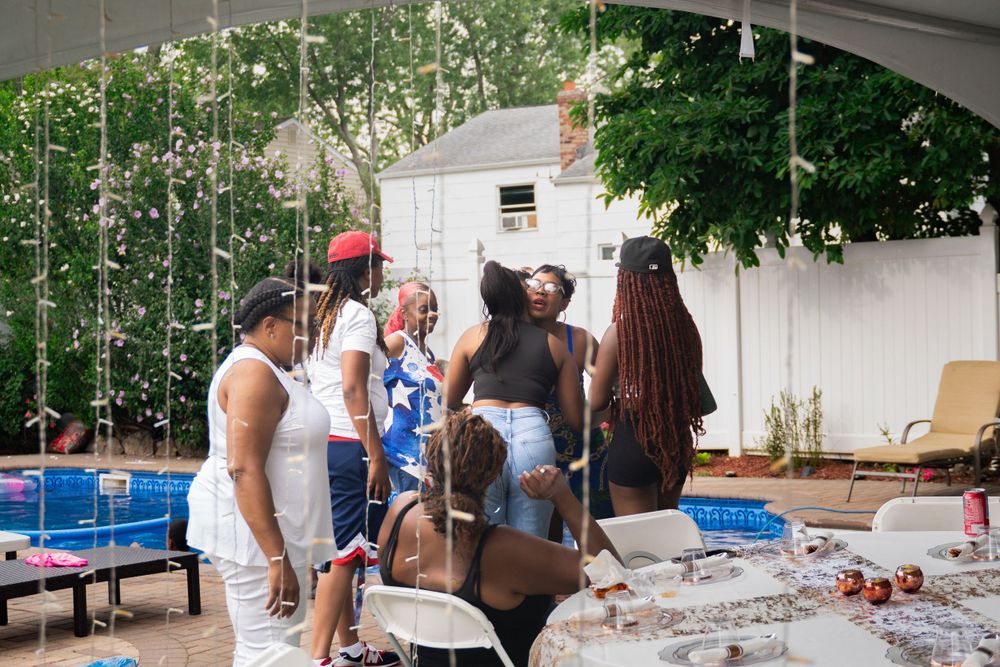 A group of people gather for a casual outdoor party with string lights and a swimming pool in the background.