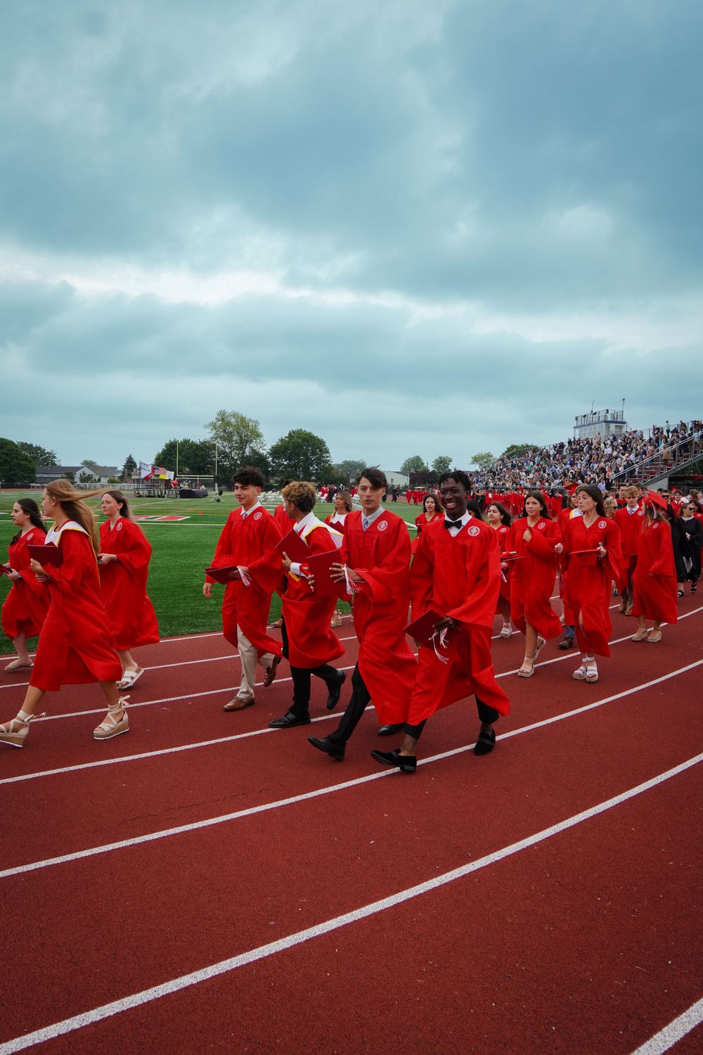 Graduates in bright red robes walk along a running track during commencement ceremony under cloudy skies.