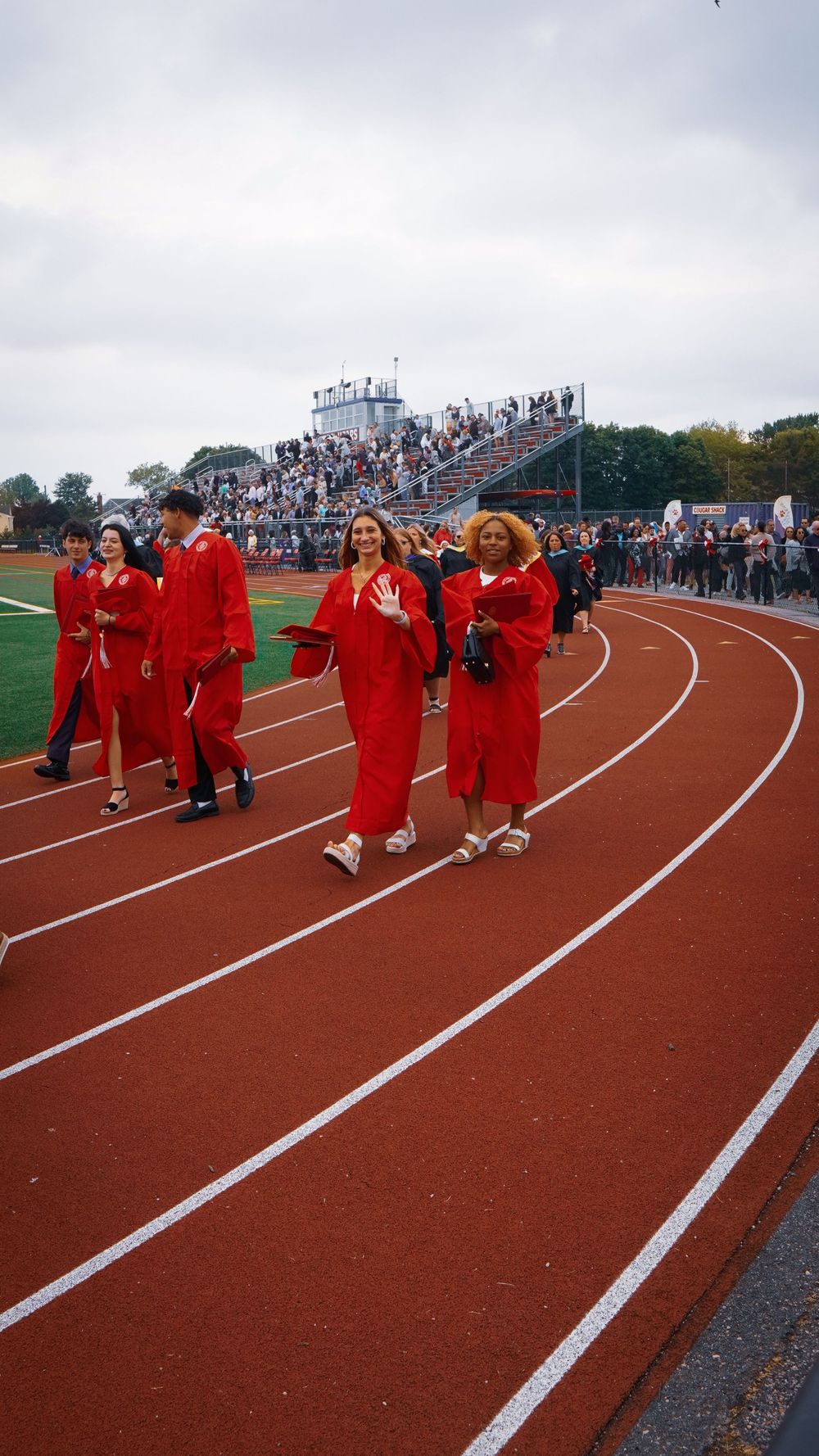 Graduates in red caps and gowns walk along a running track during an outdoor commencement ceremony with bleachers visible.