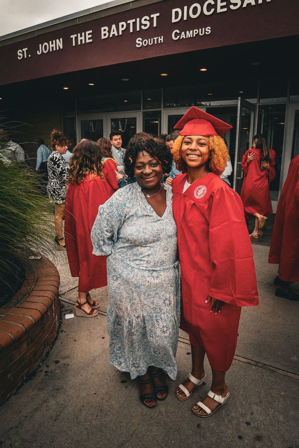 Graduation ceremony outside St. John the Baptist Diocesan South Campus with two people posing in front of the entrance.