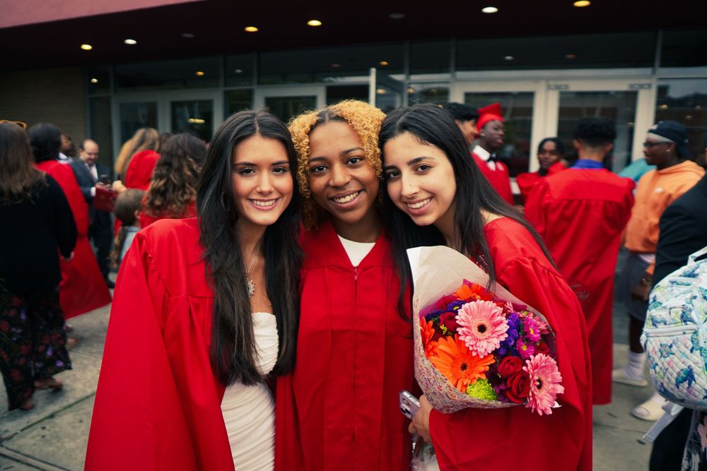Three graduates in red academic regalia celebrate together with flowers at a college commencement ceremony.