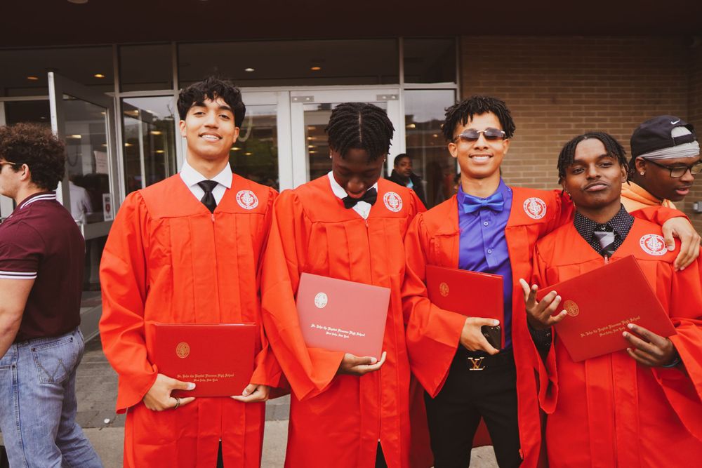 A group of students pose together wearing bright orange graduation gowns and holding diplomas outside a building.