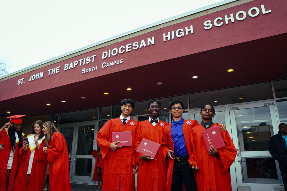 Students in bright red graduation gowns gather outside St. John the Baptist Diocesan High School entrance.