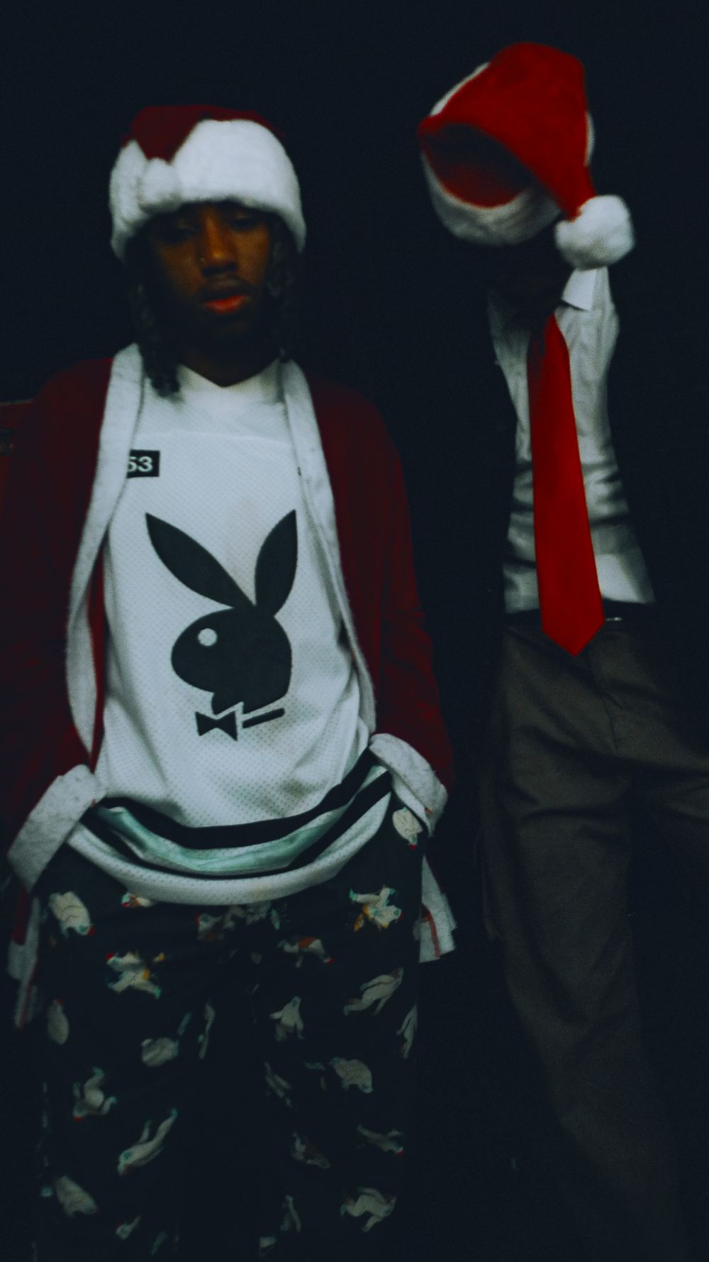 Two people wearing Santa hats and winter clothing pose against dark background, one wearing a Playboy shirt and camo pants.
