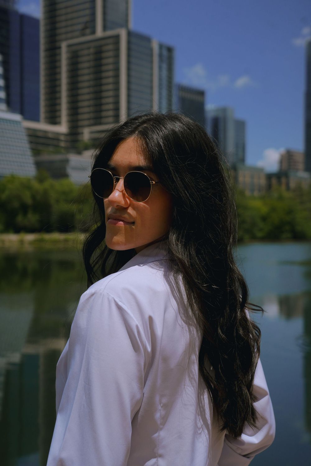Woman in white shirt and sunglasses poses near urban waterfront with skyscrapers.