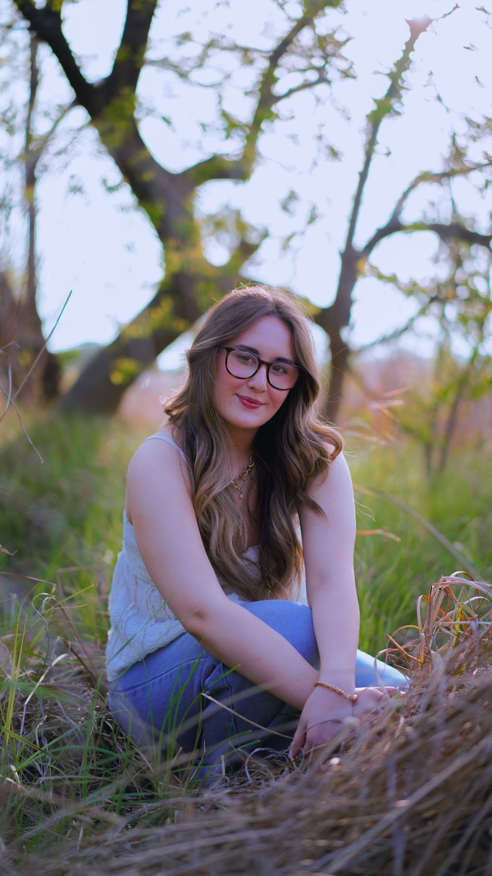 Young woman with glasses and long hair sits smiling in a grassy field with trees in the background.