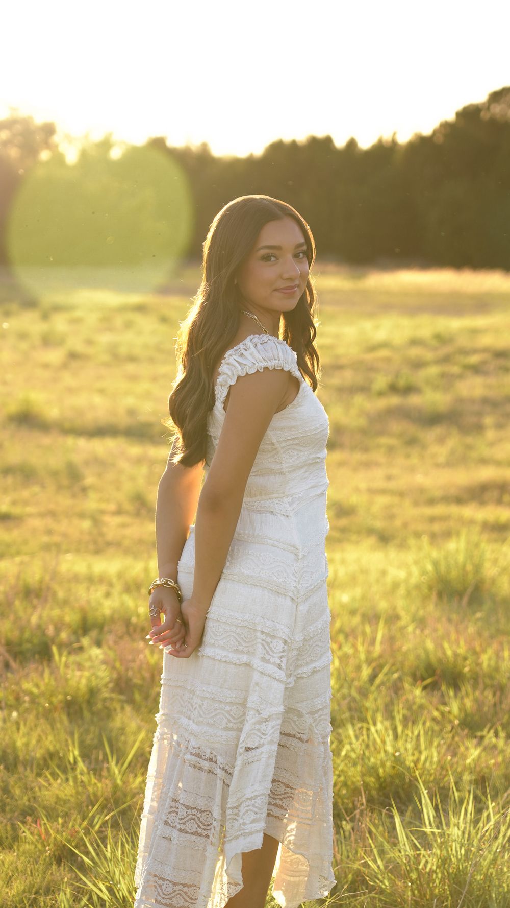 Young woman in white lace dress standing in sunlit field at golden hour, smiling over shoulder.