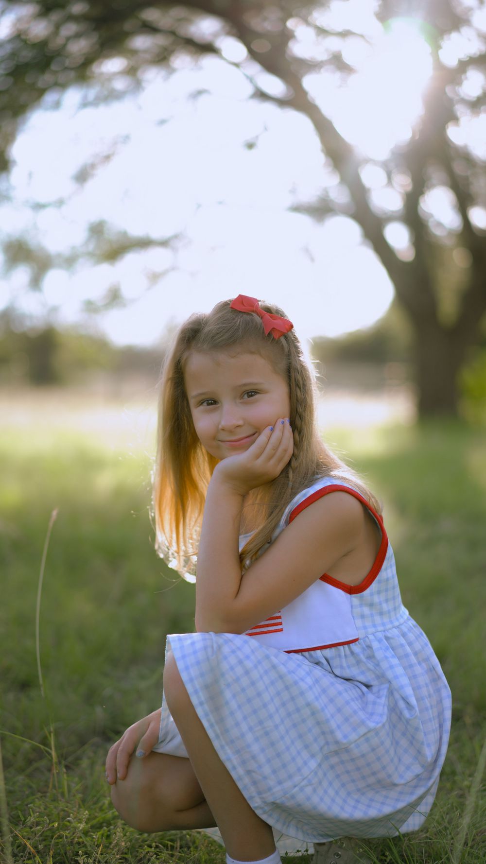 Young child in white checkered dress sitting and playing in grassy outdoor setting under natural lighting.