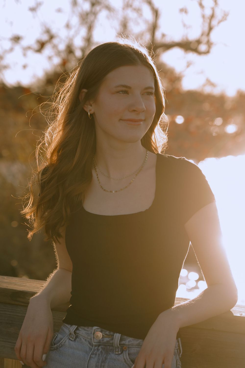 A young woman in a black top and jeans poses outdoors during golden hour with backlit sunlight creating a warm glow.