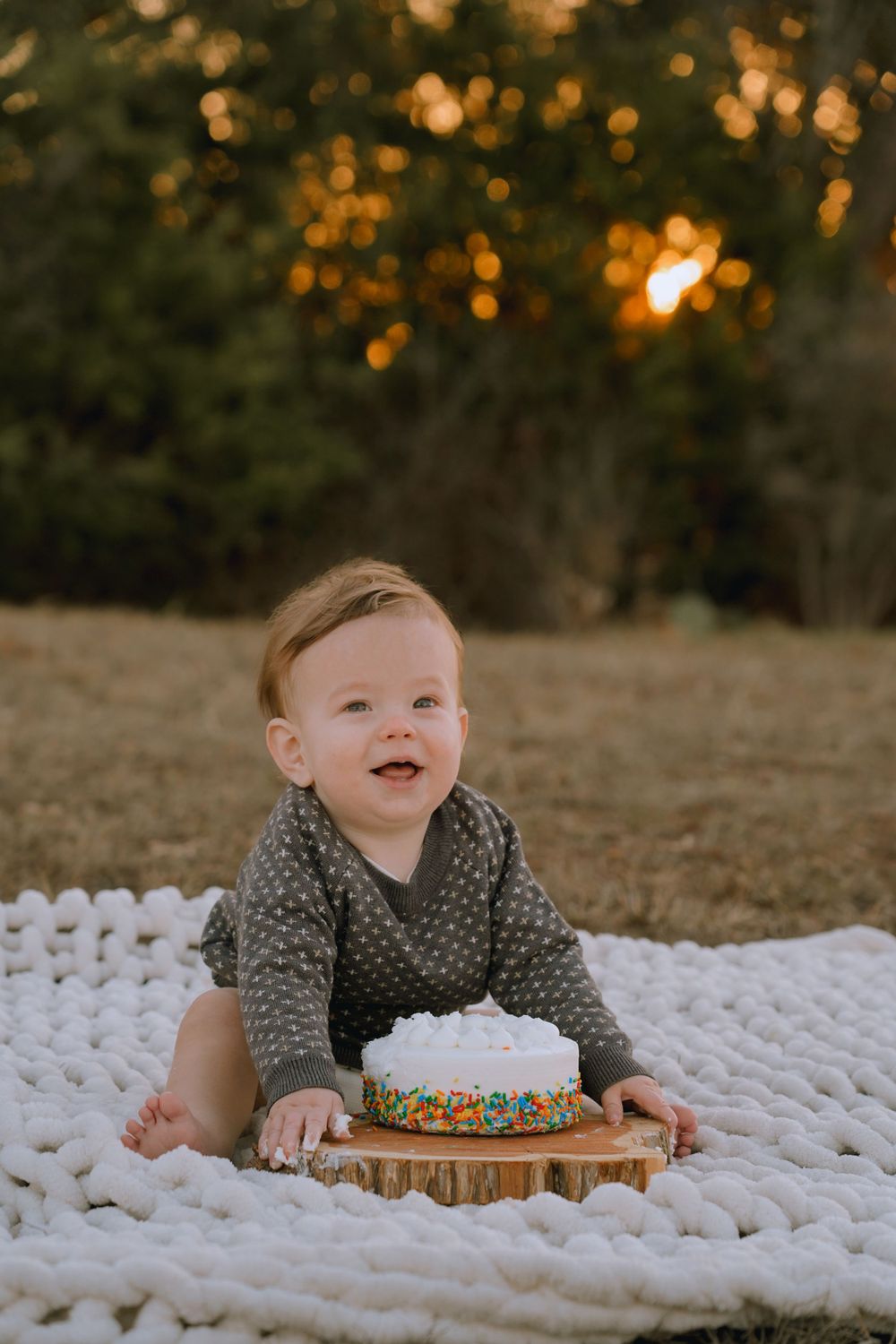 A small child wearing a grey sweater sits on a white blanket with a decorated cake at golden hour outdoors.