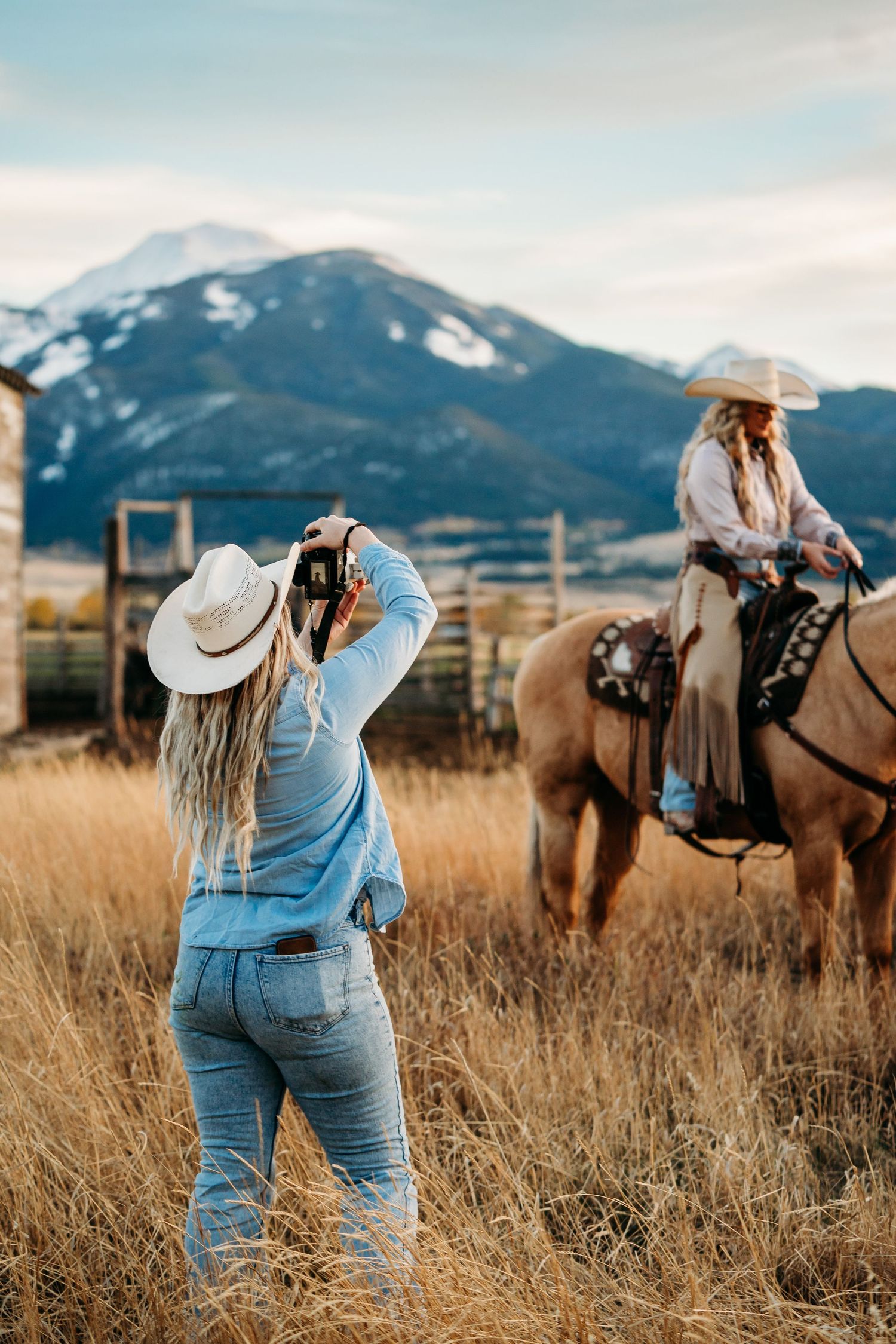 Person in denim overalls and cowboy hat photographing horse in mountain field at sunset.