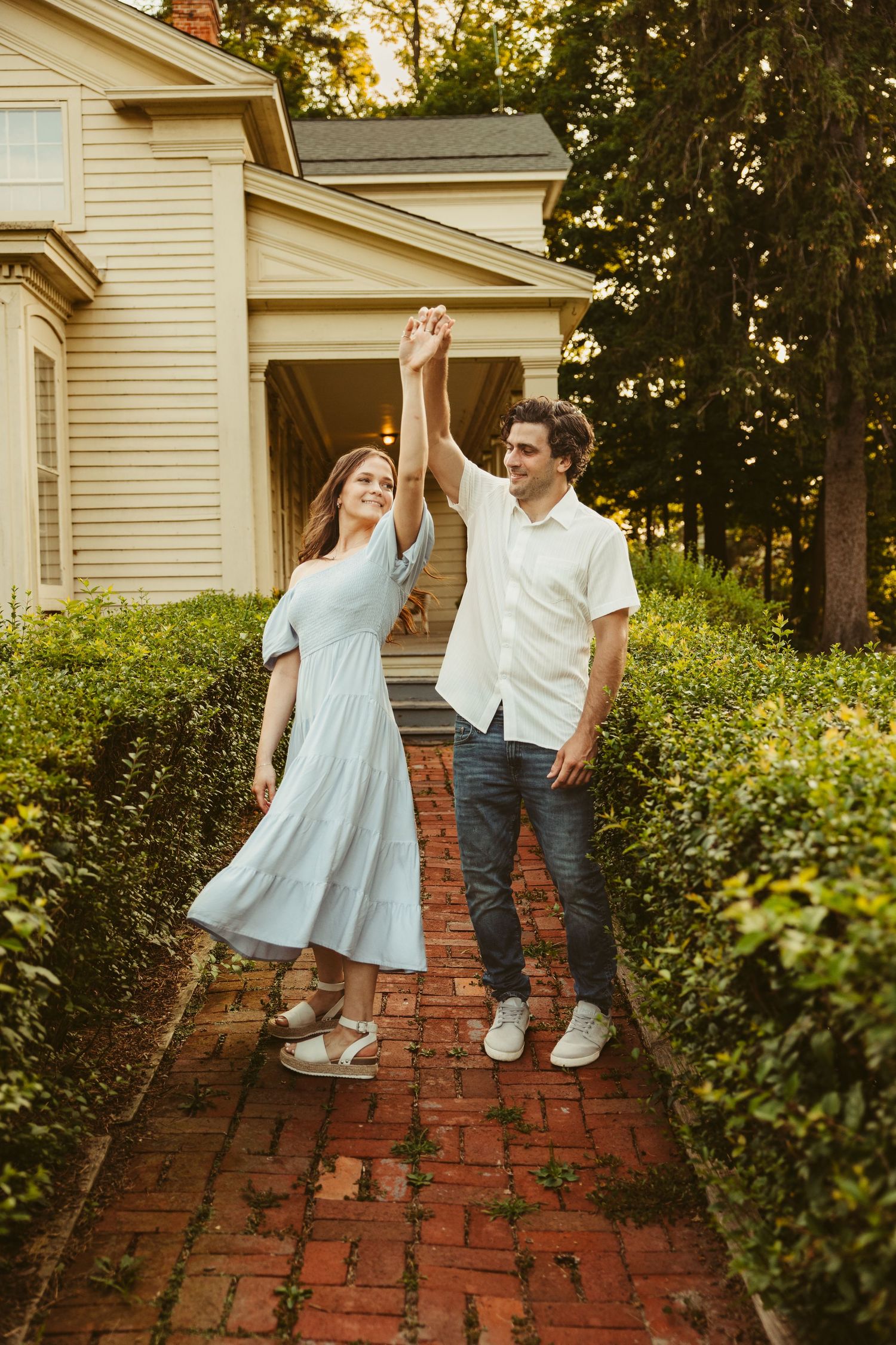 A couple dances together on a brick pathway in front of a cream-colored house during golden hour.