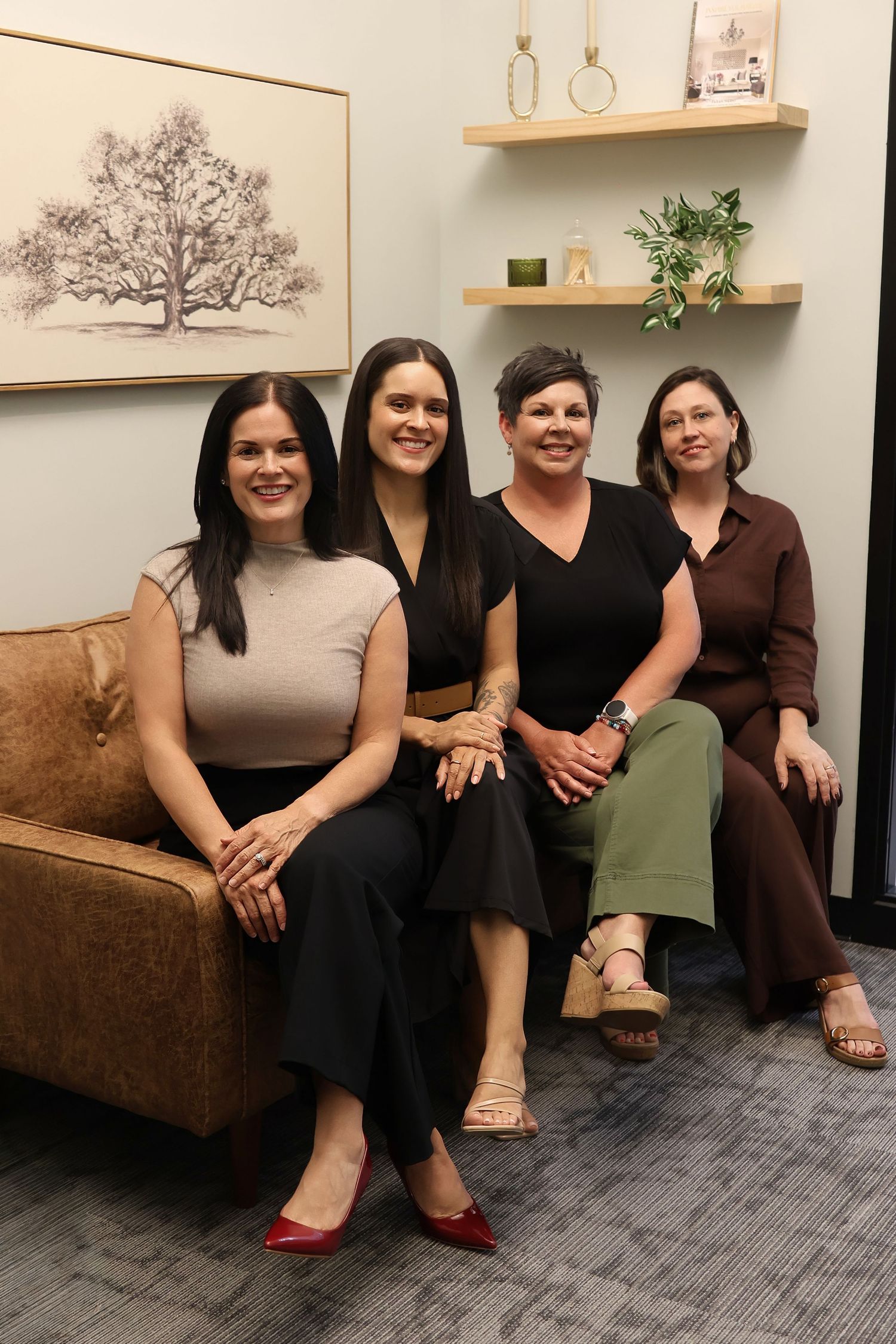 Three women in business attire seated on couch with large plant in modern office setting.