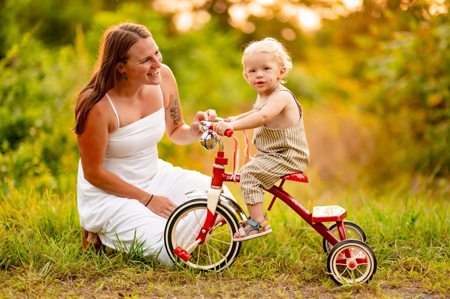 A child sits on a red tricycle in a sunny meadow while a companion kneels beside them on a warm summer evening.