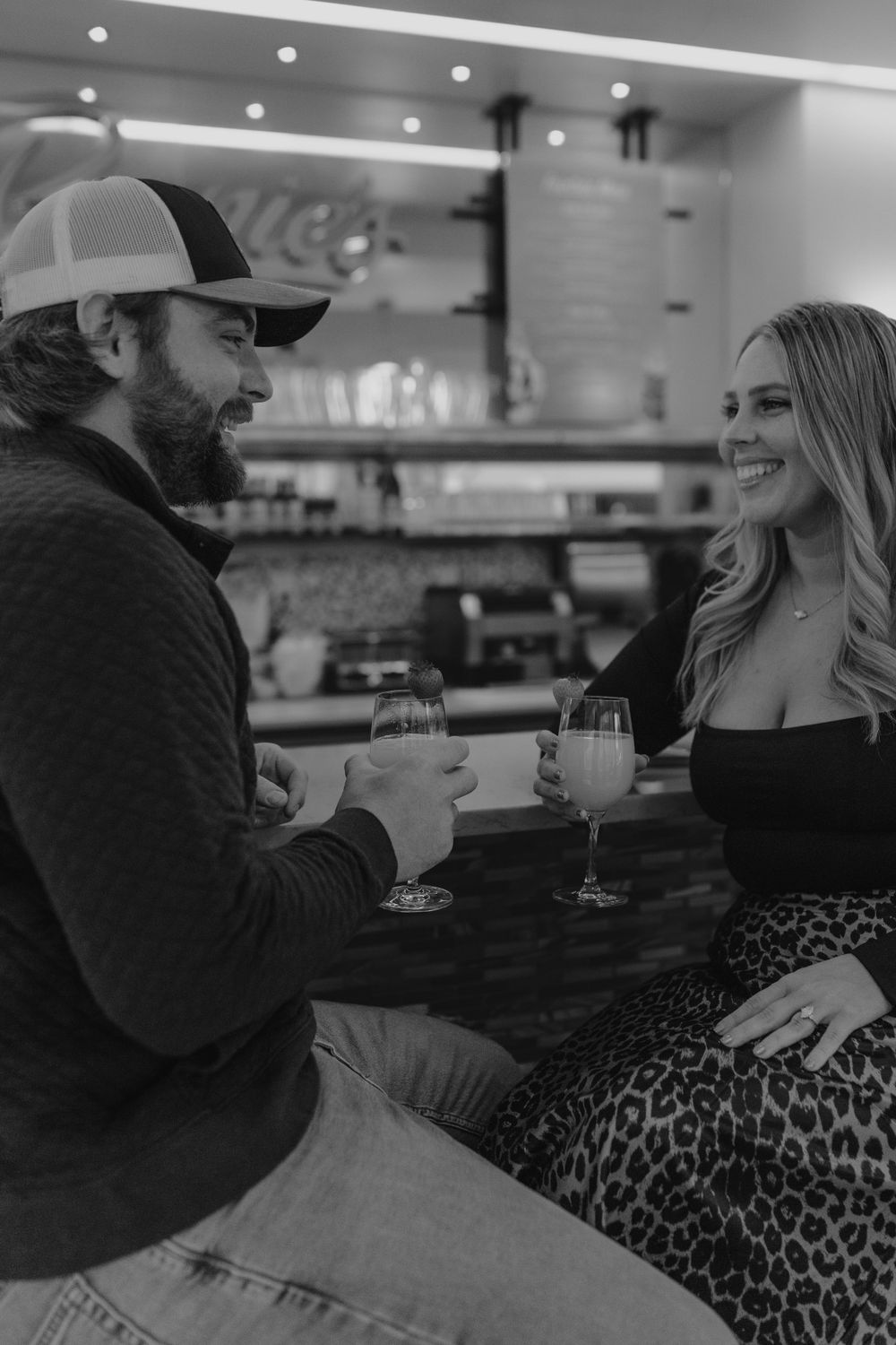 Two people share drinks at a bar counter in black and white, with one wearing a hat and the other in a leopard print skirt.