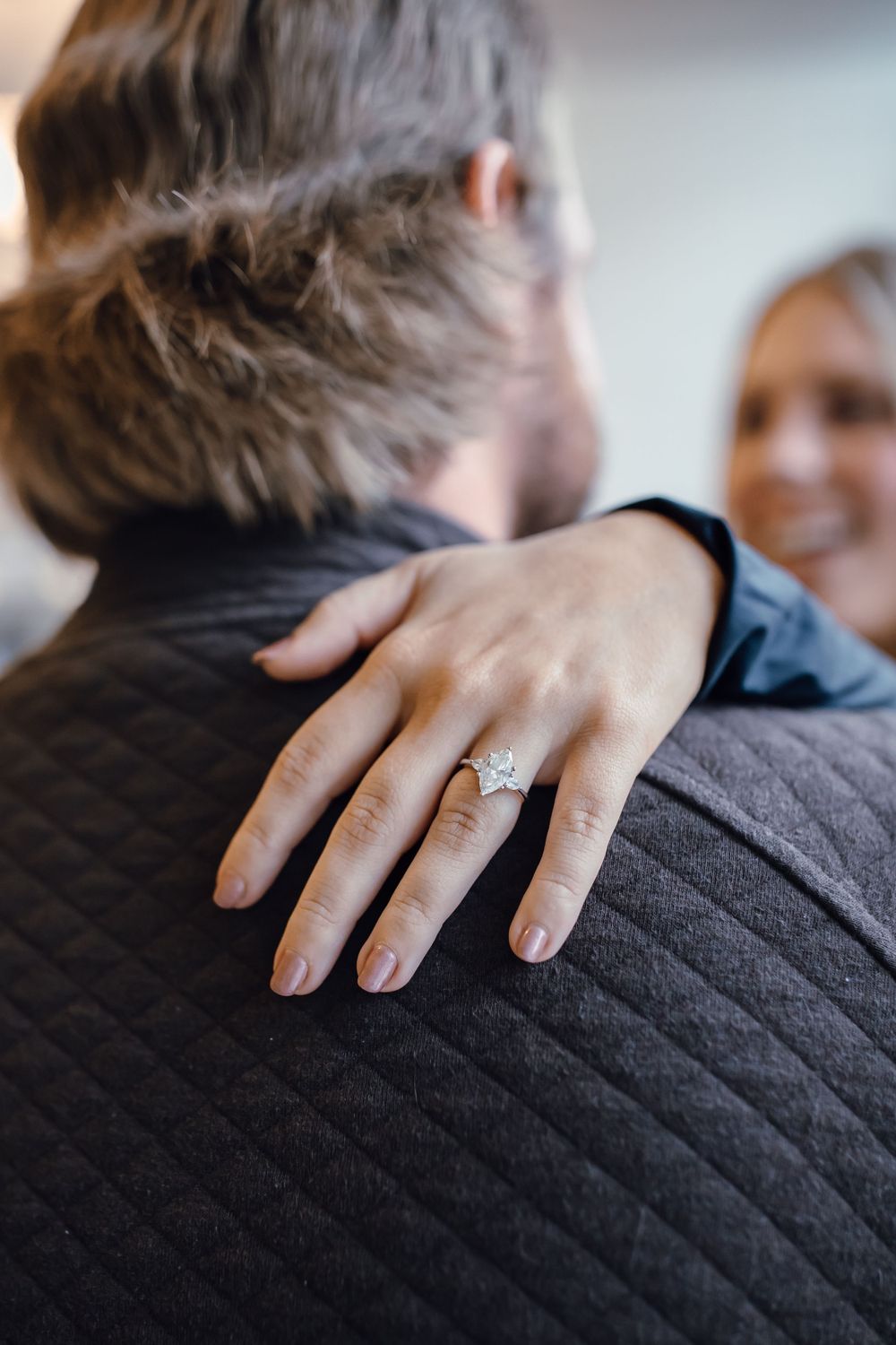 A close-up shot of an engagement ring on a hand resting on a black quilted jacket during a proposal moment.
