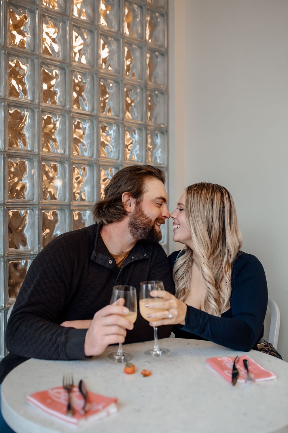 A couple sharing an intimate moment while toasting wine glasses at a table with glass block wall decor.