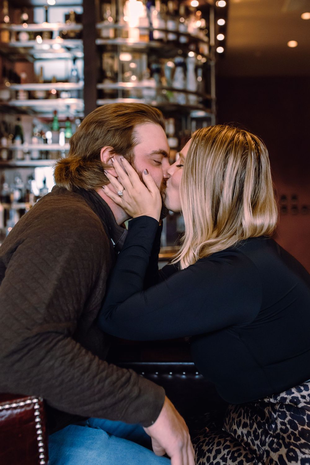 A couple shares a romantic kiss at a cozy bar with shelves of bottles in the background.