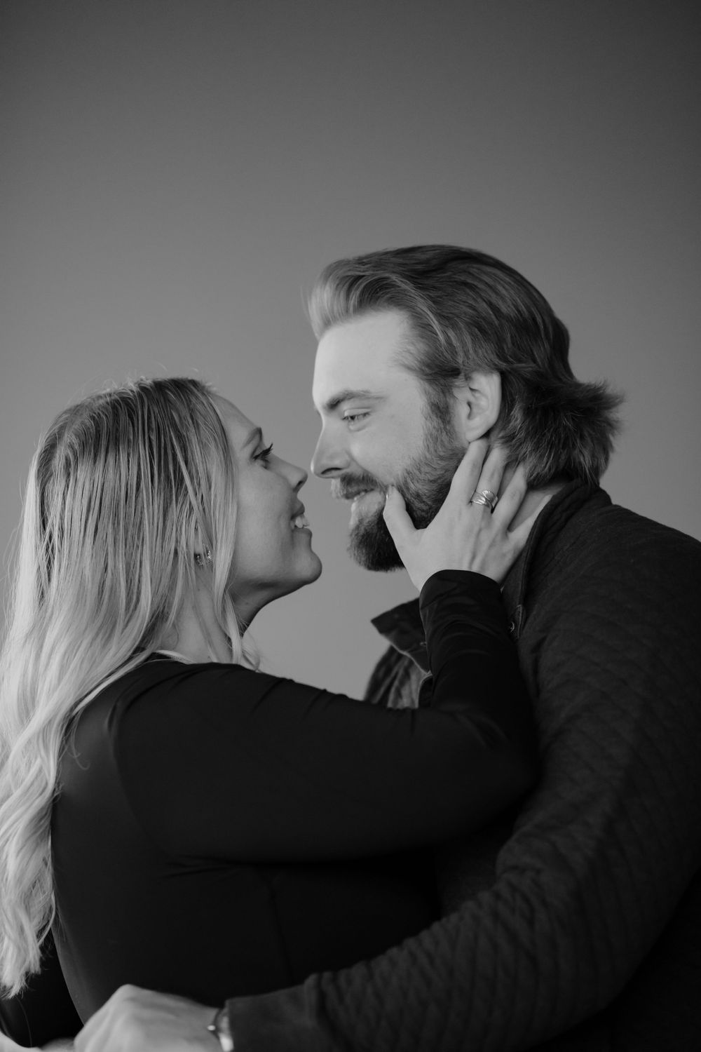 Intimate black and white portrait of a couple sharing a tender romantic moment against simple grey background.