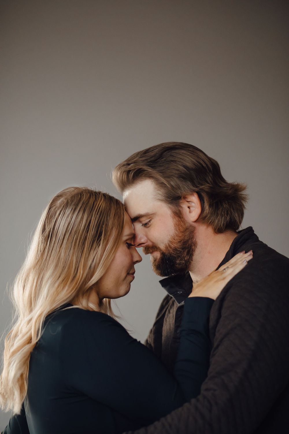 An intimate moment between a couple in dark clothing as they embrace with foreheads touching against a neutral background.