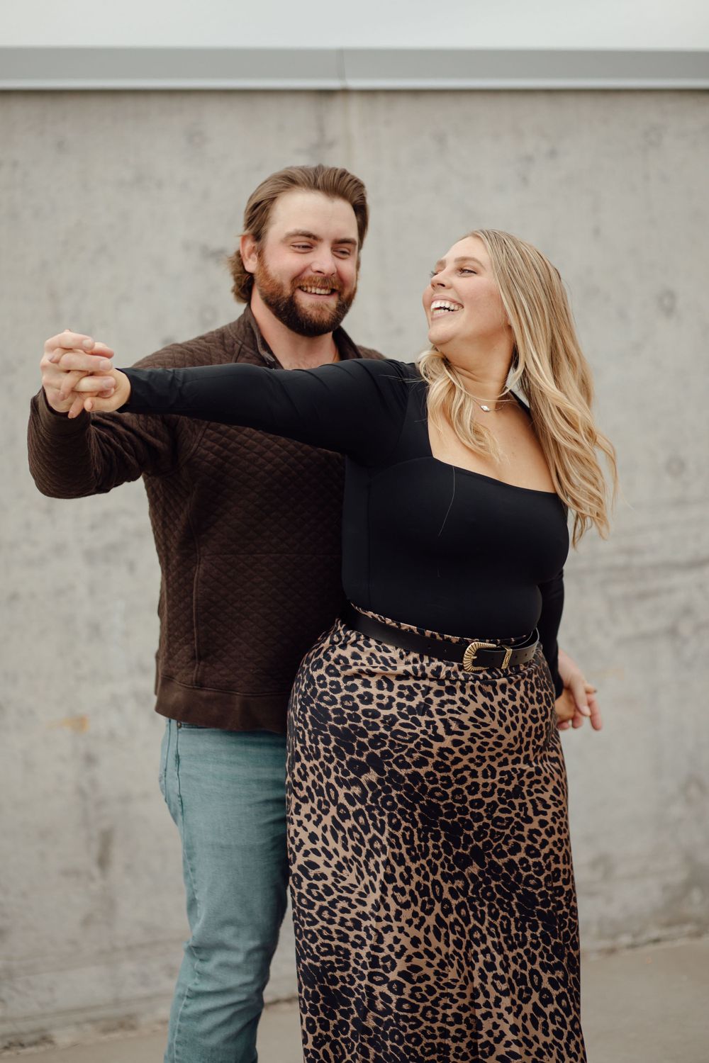 A couple laughs and dances together, with her wearing a leopard print skirt and him in a brown sweater near a gray wall.