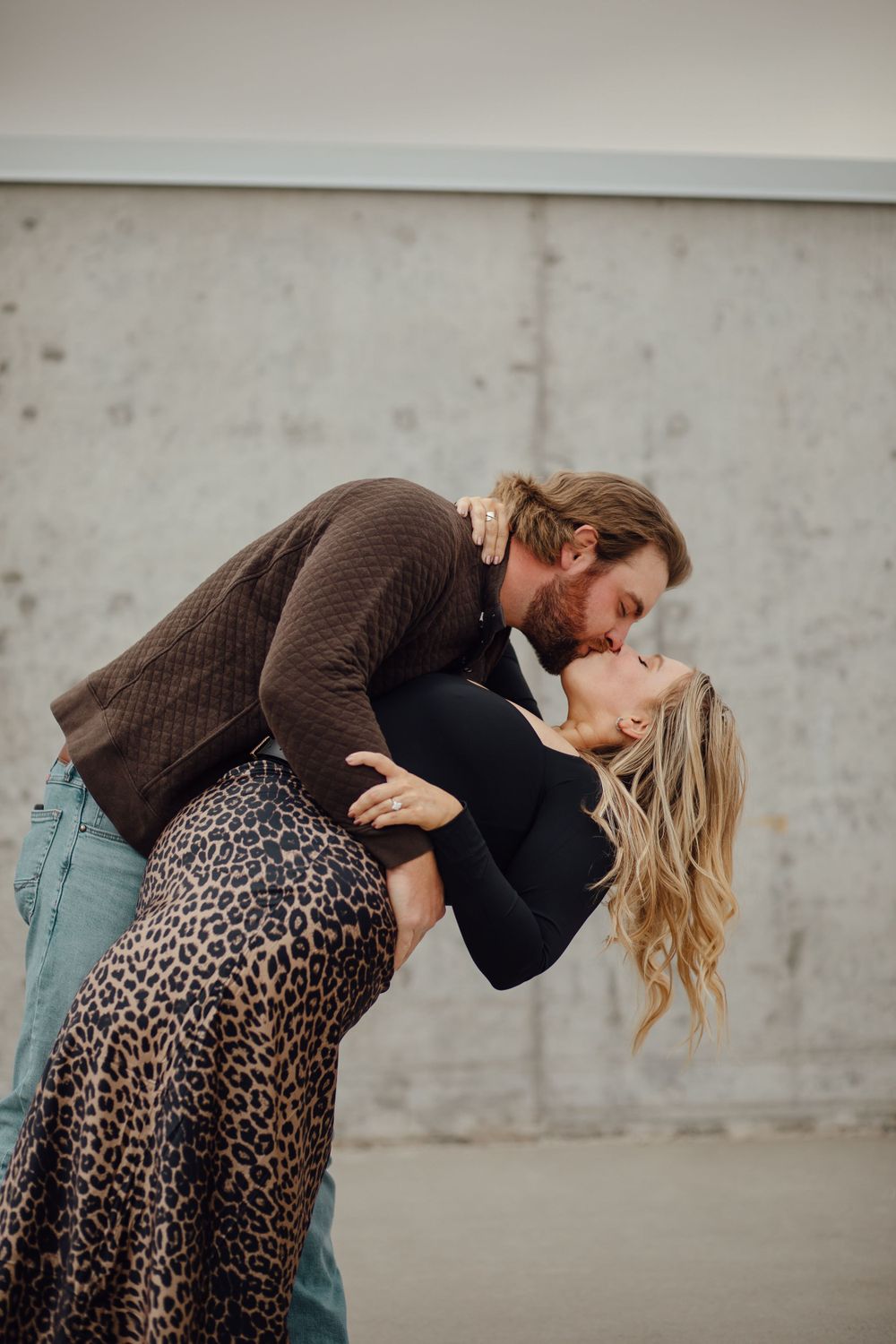 A romantic couple in a candid pose, with her wearing a leopard print skirt and him in a brown sweater against a concrete wall.