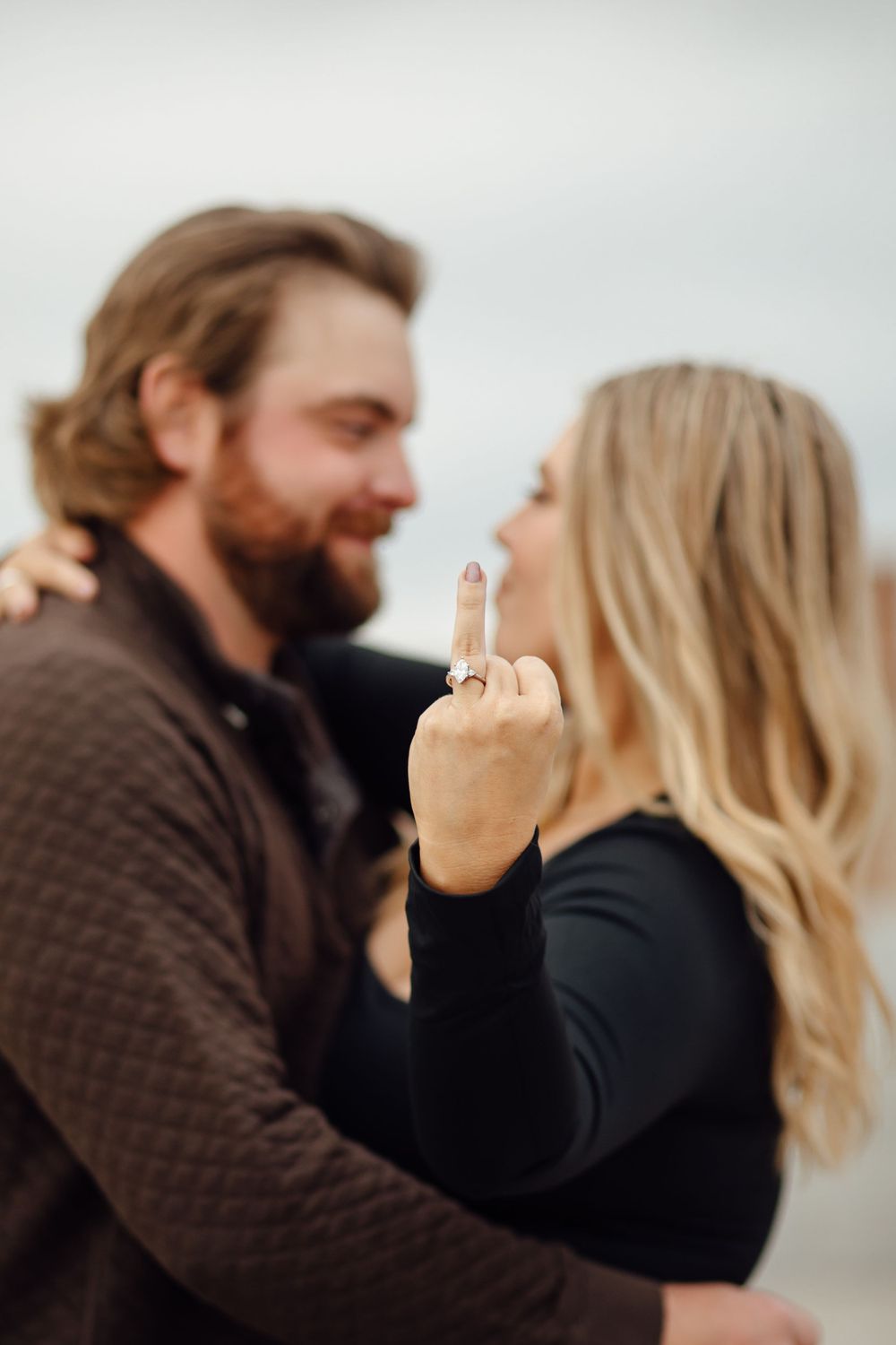 A couple in dark clothing embraces while the woman playfully shows her middle finger to the camera.