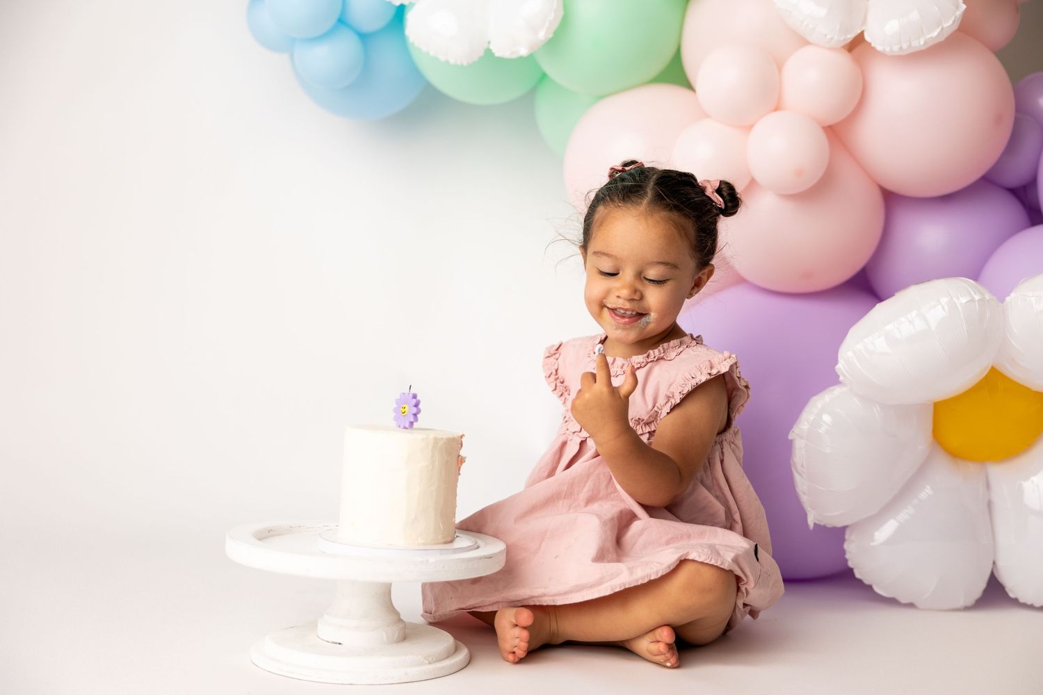 A young child in a pink dress sits beside pastel balloon decorations and a birthday cake on a white platform.