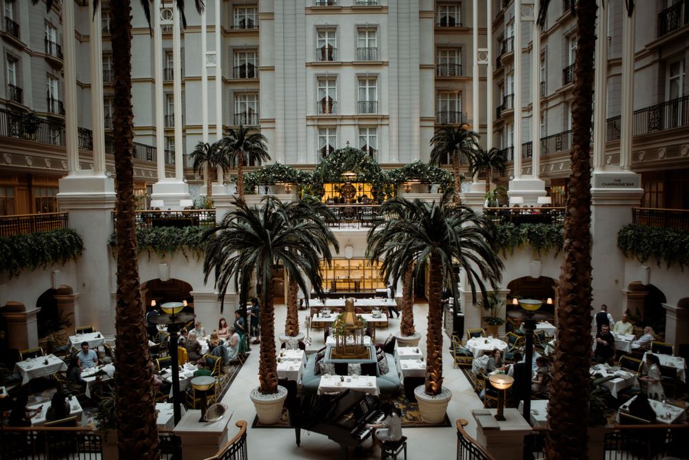 Luxurious hotel atrium with palm trees and elegant dining tables in an opulent European-style interior setting.
