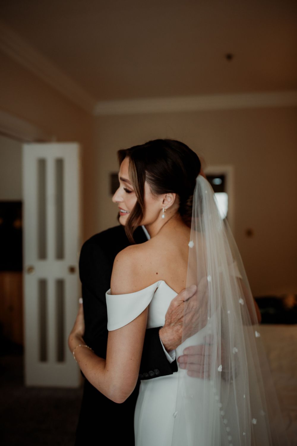 A bride in an elegant off-shoulder white wedding gown embraces someone in black during an intimate moment indoors.