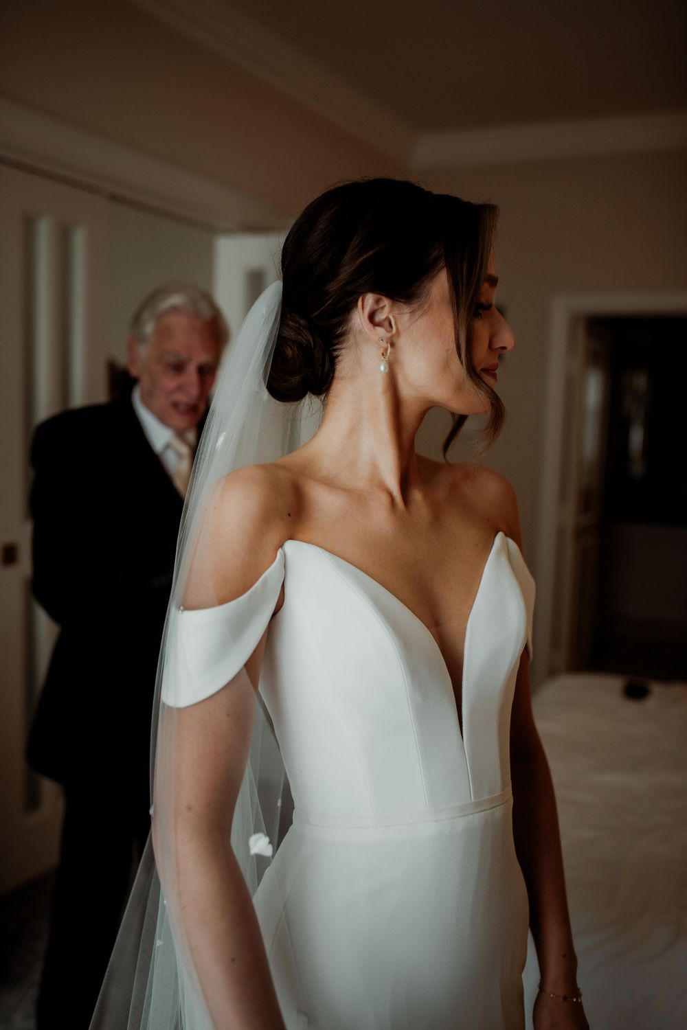A bride in an elegant off-the-shoulder white wedding dress with a contemporary design stands in a dimly lit room.