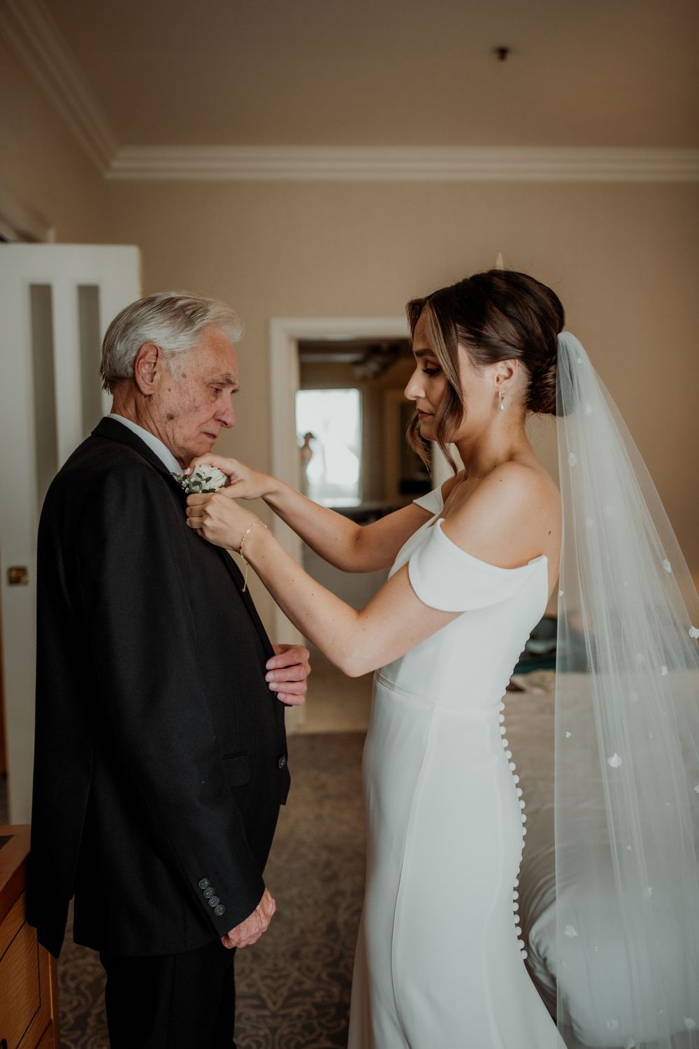 Tender moment showing a bride in white dress and veil adjusting a boutonniere on an older gentleman's suit lapel.