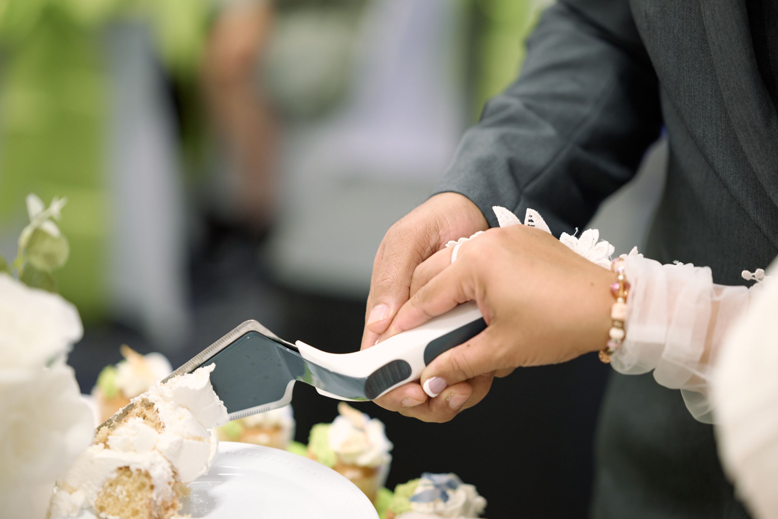 Close-up of hands cutting a white wedding cake with ivory flowers at an outdoor reception at Fairfield Inn & Suites Lodi, California.