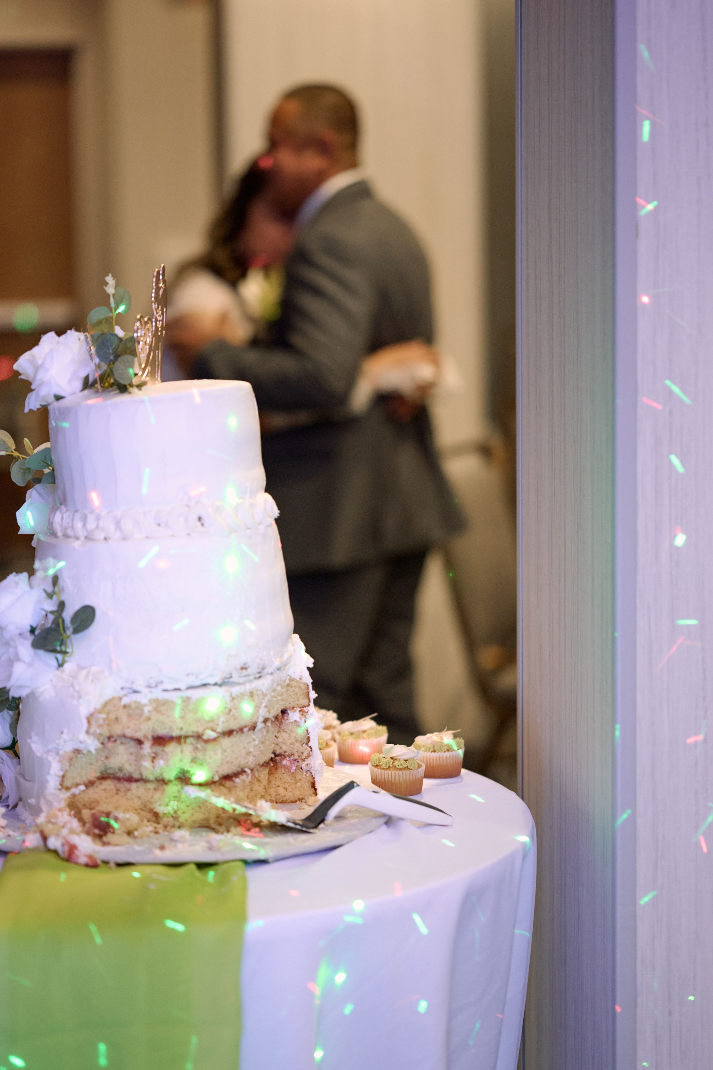 Elegant white wedding cake on display with green lighting while a couple embraces in the blurred background at Fairfield Inn & Suites Lodi.