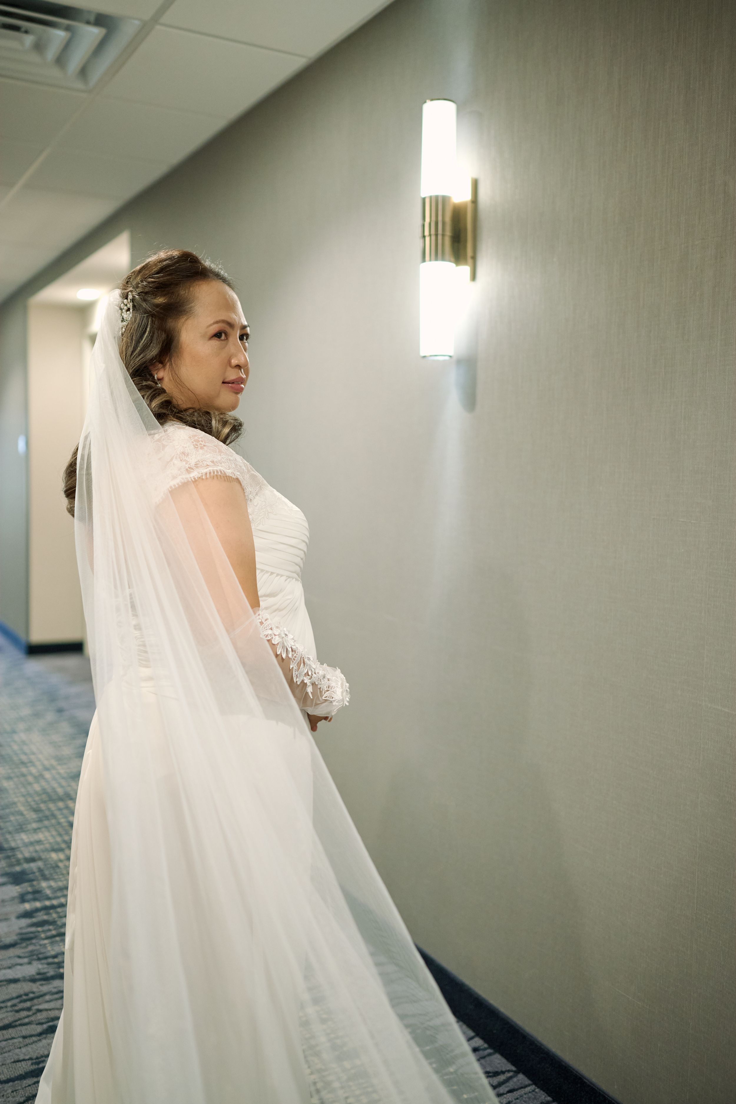 A bride in a white wedding dress and veil stands in a hallway with modern wall sconce lighting at Fairfield Inn & Suites Lodi, California.