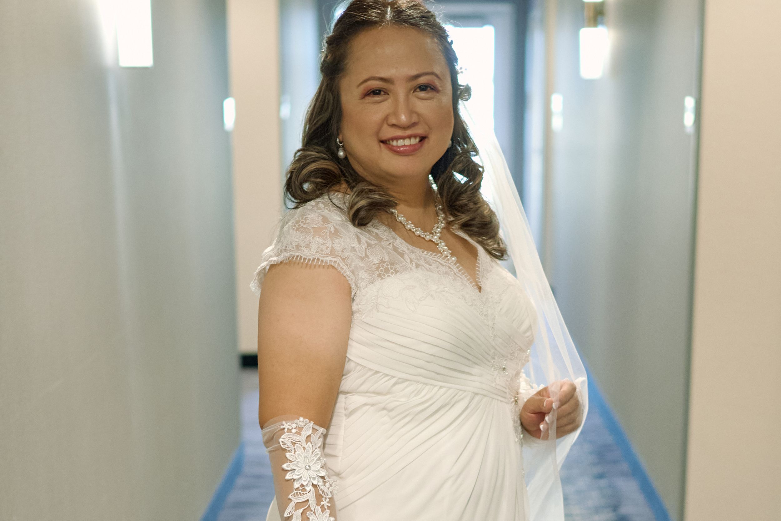 A bride in a white lace wedding dress with cap sleeves and lace gloves stands smiling in a hallway at Fairfield Inn & Suites Lodi, CA.
