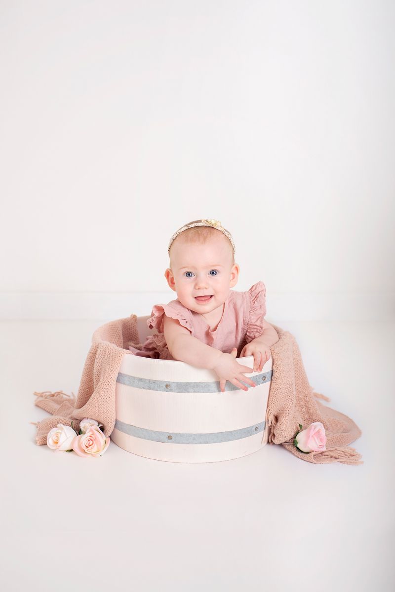 6 month old sits in white wooden prop with pink romper and pink roses