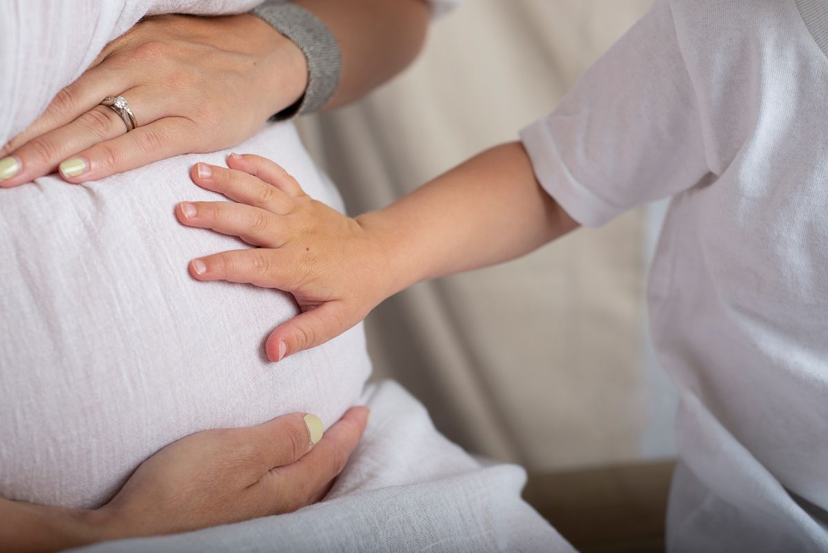 close up of mom in simple white dress and young child places little hand on moms pregnant belly for in studio maternity session*