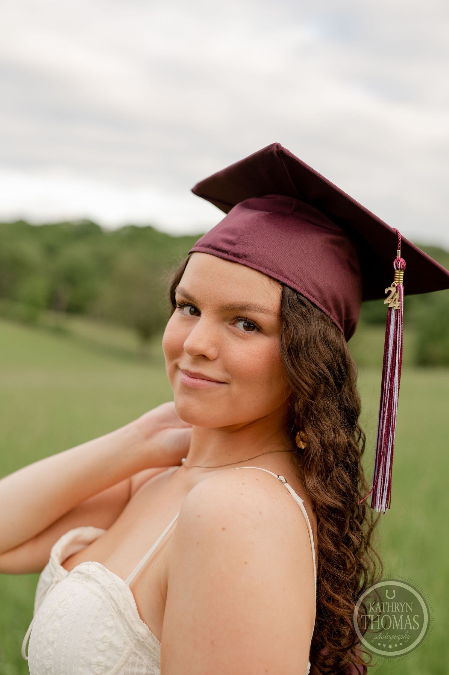 Graduate in a burgundy cap and white dress poses for a portrait in a sunny meadow.