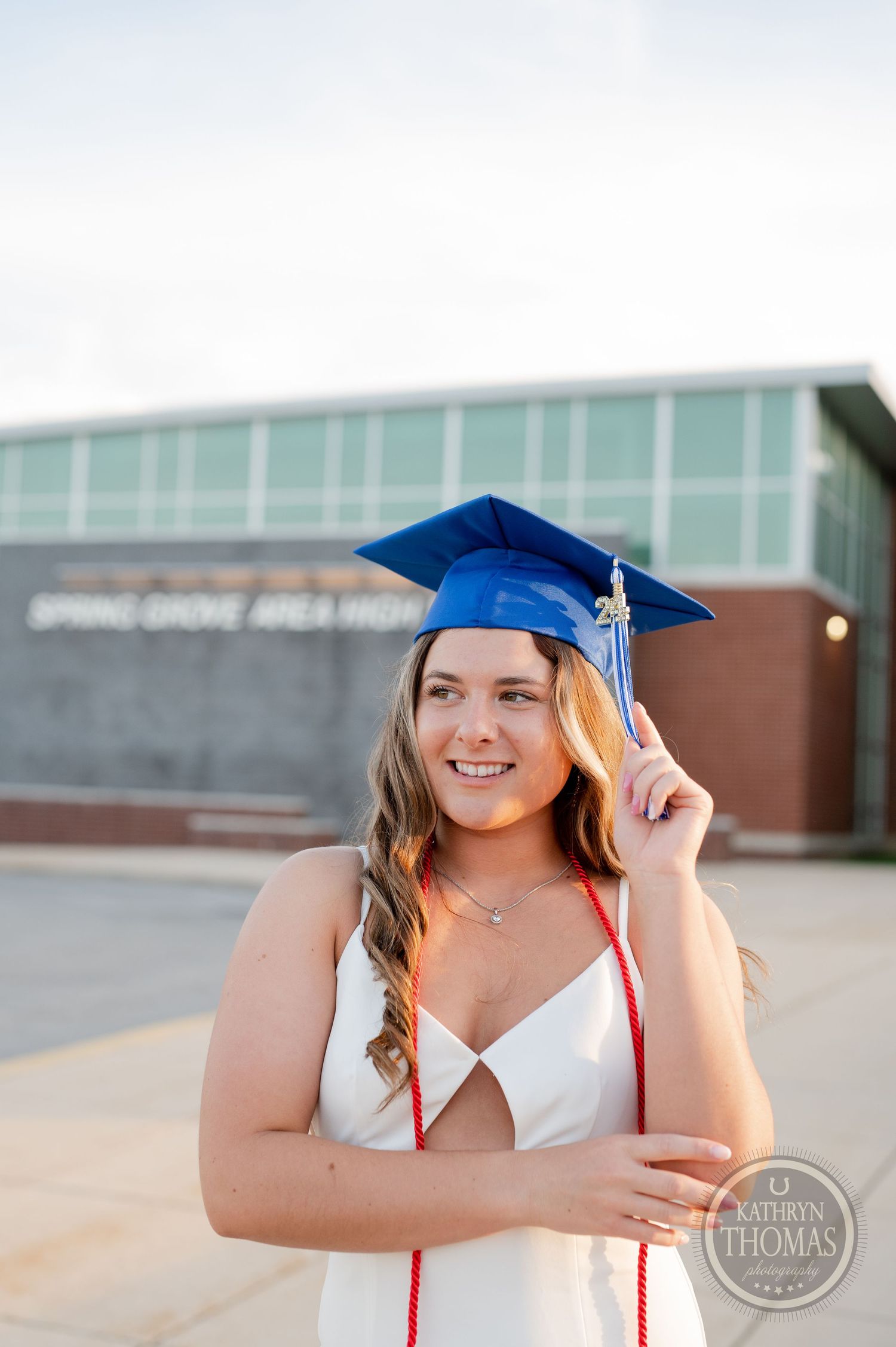 Graduate standing outside a school building wearing white dress and blue cap while holding tassel.