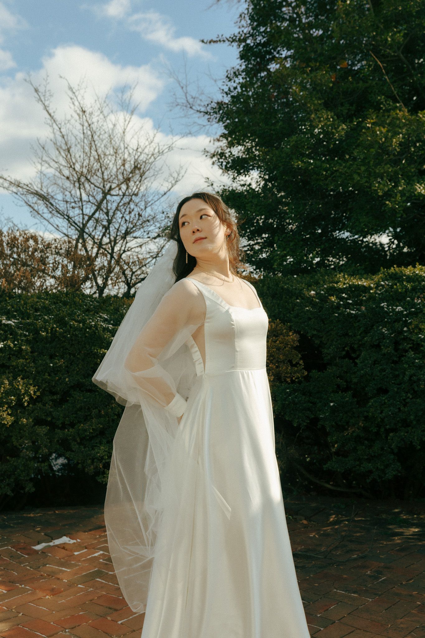 A bride in a flowing white wedding gown with veil poses outdoors against greenery and a blue sky on a sunny day.