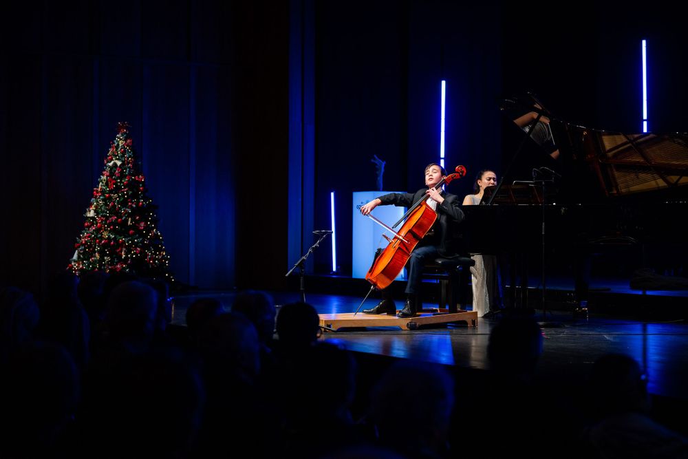 Classical music performance featuring a cellist and piano on a darkly lit stage with a Christmas tree in background.