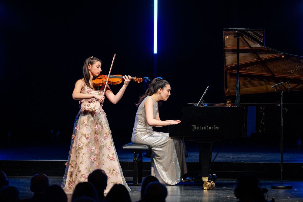 Duo performance featuring violinist in flowing dress and pianist at grand piano on concert stage.