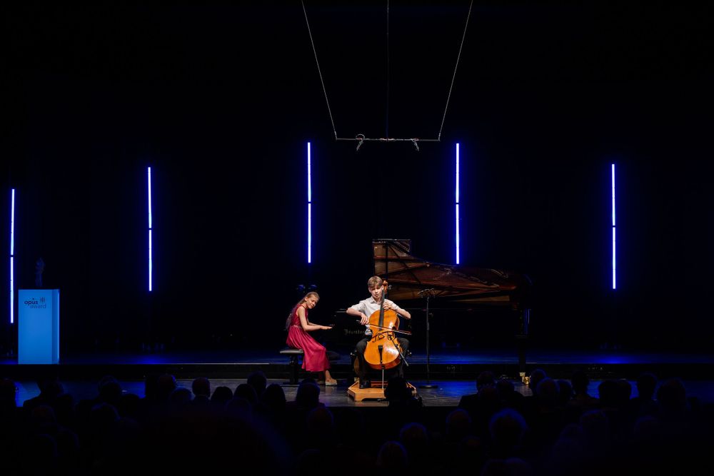 Duet performance with cellist and pianist under striking blue stage lighting.