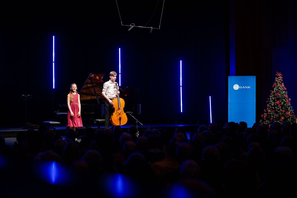 Concert performance with cello player near decorated Christmas tree and blue vertical light installations.