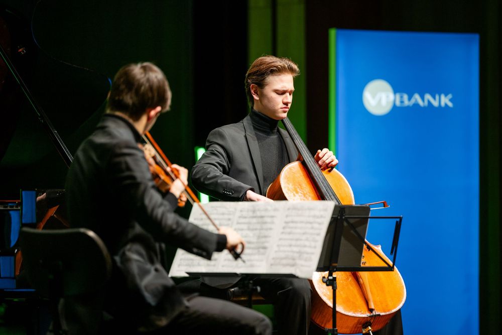 Two classical musicians perform with string instruments against a blue backdrop.